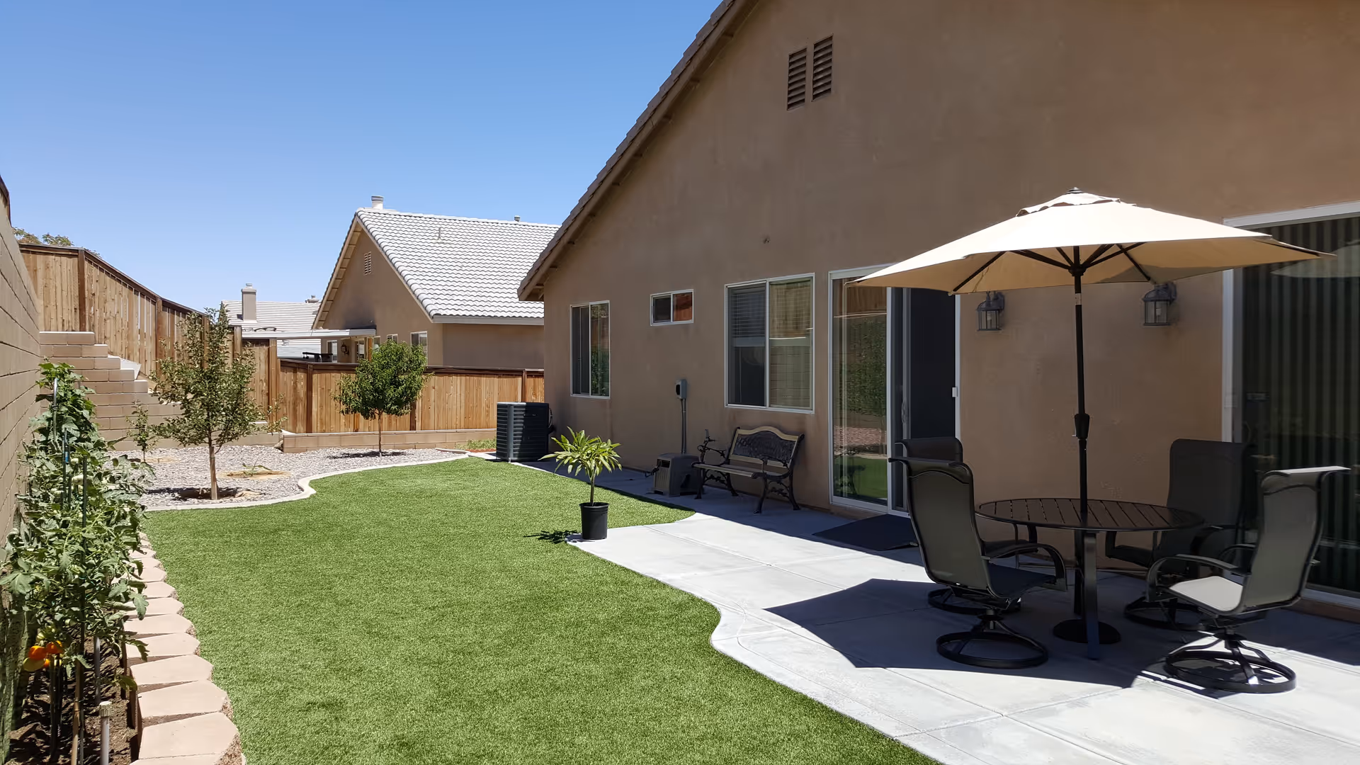 Backyard area of a residential facility with artificial grass, small trees, and a concrete patio. The patio has a round table with four chairs and a large beige umbrella. The building has beige walls and several windows and sliding glass doors. A wooden fence encloses the yard, and there are steps leading up on the left side.