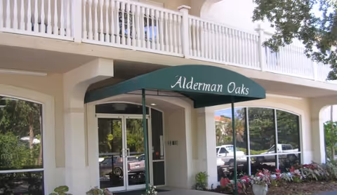 Entrance of Alderman Oaks Retirement Residence featuring a green awning with the facility name above glass double doors, surrounded by beige building walls and large windows, with plants and trees nearby.