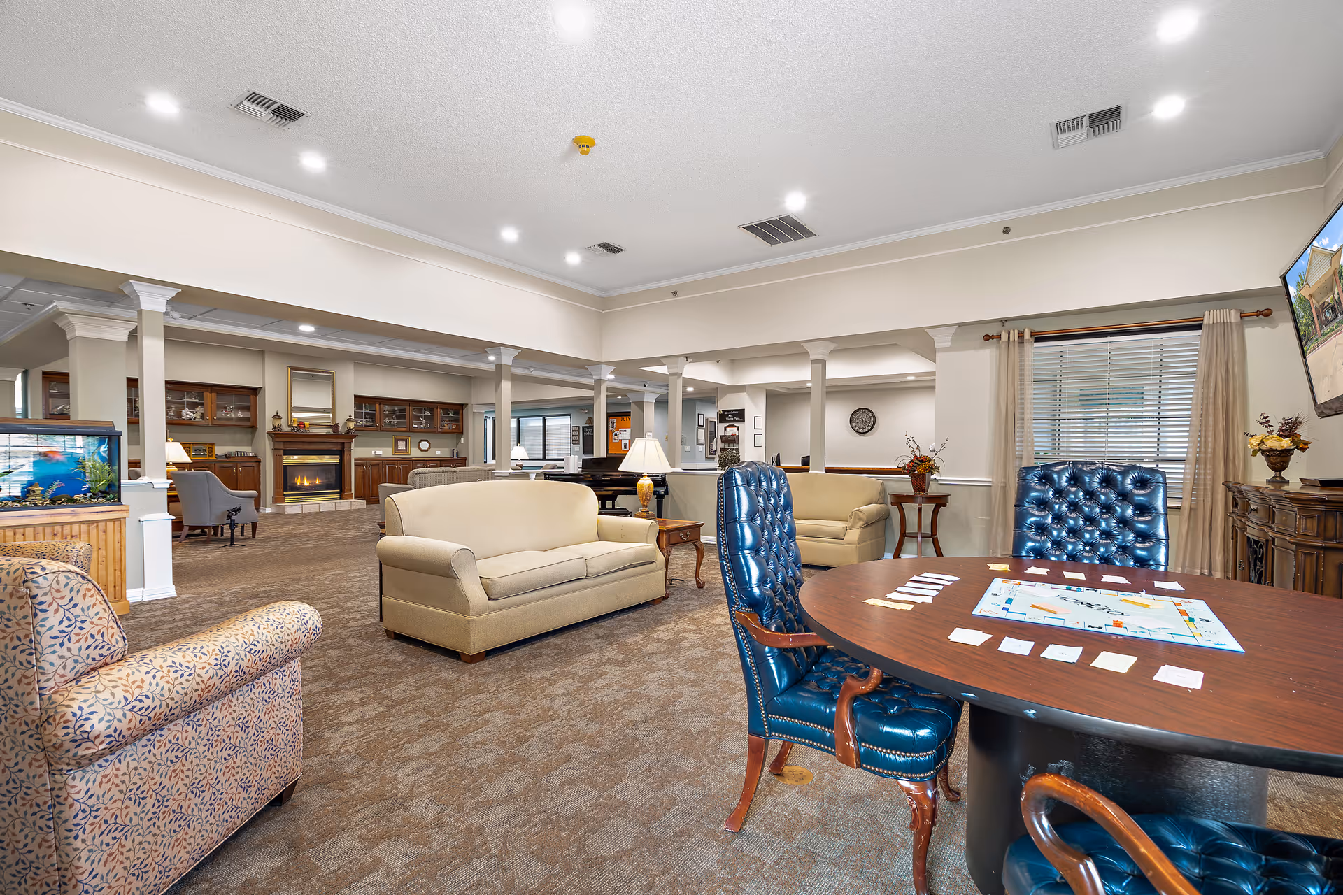 A spacious senior living facility common area with comfortable seating including beige sofas and blue leather chairs around a wooden table with a board game. The room features carpeted floors, a fireplace with a mantle, built-in wooden cabinets, a fish tank, and large windows with curtains allowing natural light.