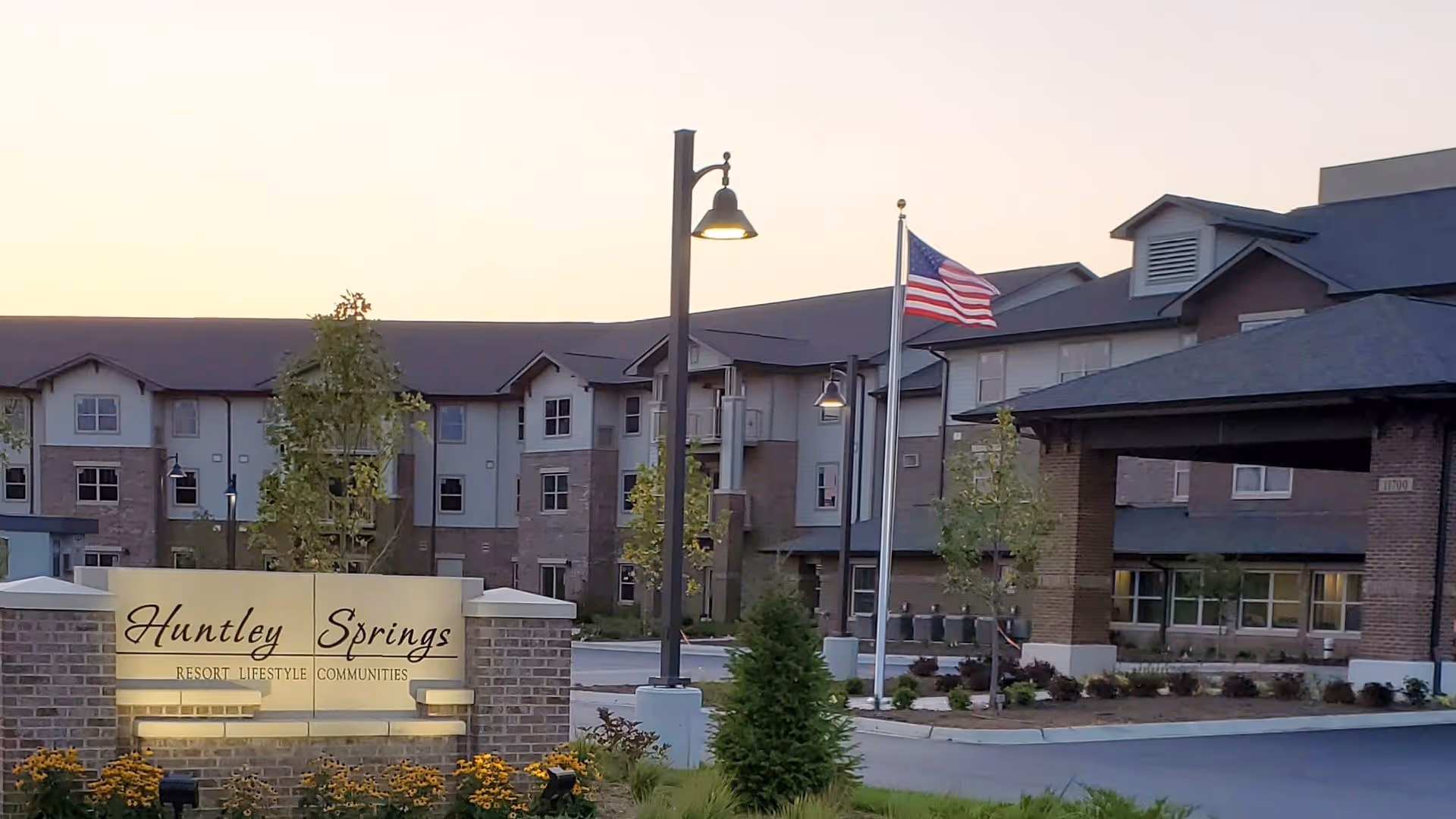Exterior view of Huntley Springs Retirement Resort at dusk, showing a large multi-story building with a covered entrance, an American flag on a flagpole, street lamps, and a landscaped area with flowers and trees. A sign in front reads 'Huntley Springs Resort Lifestyle Communities.'