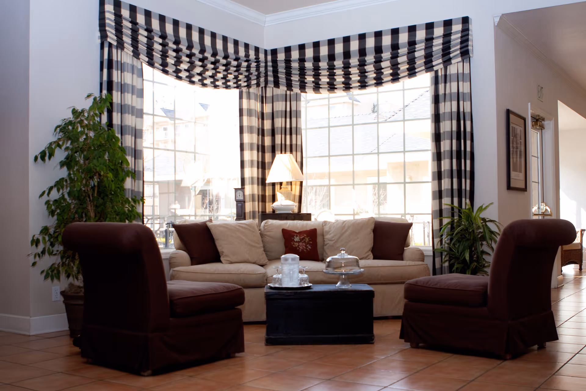 A cozy living room area with a beige sofa adorned with multiple cushions, two dark brown armless chairs, a black coffee table with a glass cake stand and water pitcher, large windows with black and white checkered curtains, and potted plants on either side of the sofa.