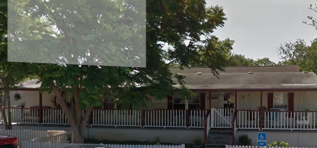 Exterior view of a single-story building with a covered porch and railing, surrounded by trees and a white picket fence. There is a handicap parking sign visible near the entrance steps.