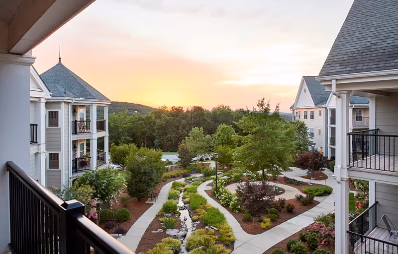 View of a landscaped garden courtyard at sunset, surrounded by multi-story residential buildings with balconies. The garden features winding pathways, a small stream, various trees, shrubs, and flower beds.