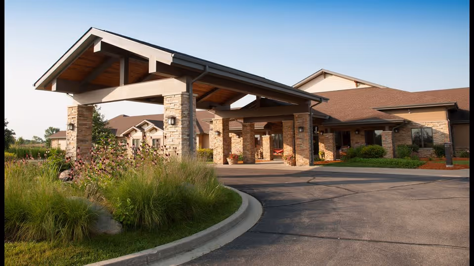 Exterior view of NorthPointe Terrace facility showing a covered entrance with stone pillars, a driveway, and landscaped greenery with tall grasses and flowering plants under a clear blue sky.
