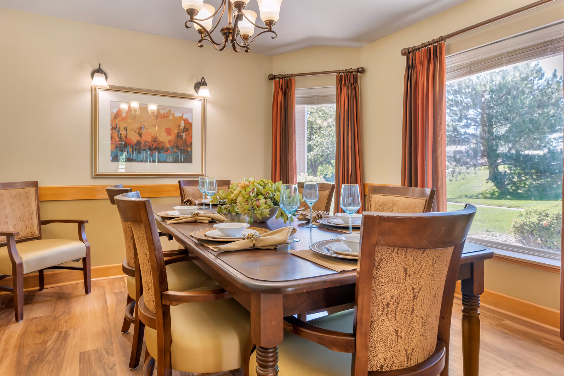 Dining room with a wooden table set for six, upholstered chairs, chandelier, wall art, and large windows overlooking a landscaped yard.