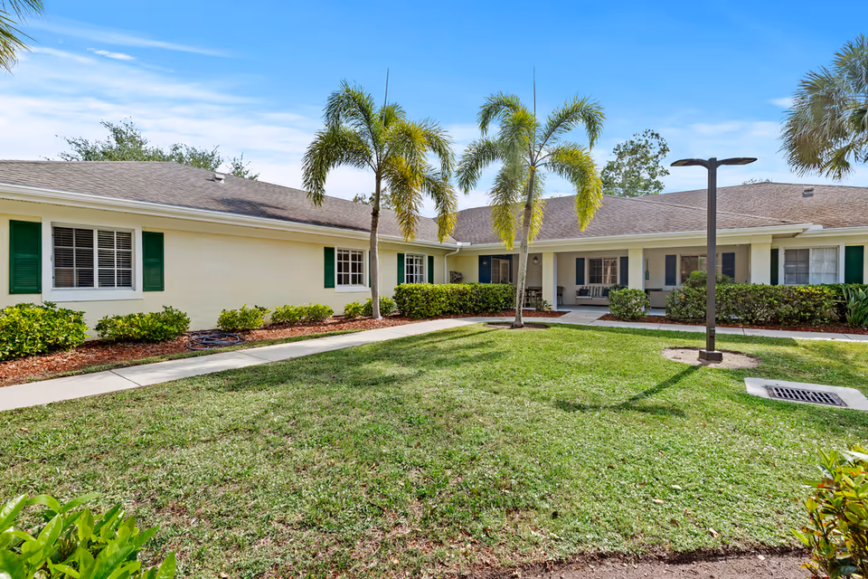 Exterior view of a single-story senior living facility building with beige walls, green window shutters, and a brown shingled roof. The building surrounds a grassy courtyard with palm trees, bushes, a sidewalk, and a lamp post under a blue sky with some clouds.