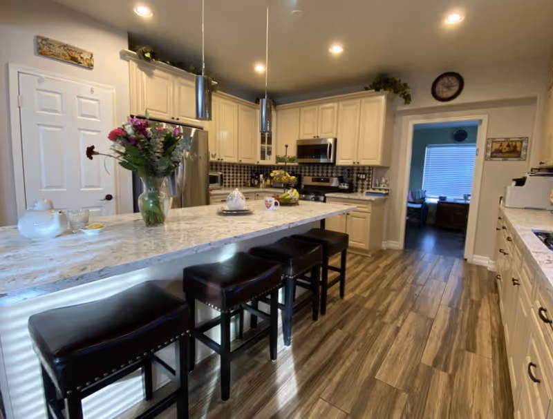 A modern kitchen with a long marble countertop island featuring four dark leather stools. The kitchen has cream-colored cabinets, stainless steel appliances including a refrigerator and microwave, and a tiled backsplash. There is a vase with flowers on the island, and the floor is covered with wood-patterned tiles. A doorway leads to another room with a window and blinds.