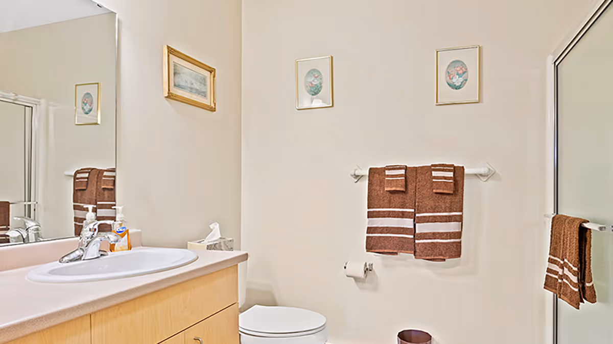 A clean bathroom with a light wood vanity and white sink, a large mirror above the sink, a toilet, a glass shower door with a brown towel hanging on the handle, two brown towels with white stripes hanging on a towel rack above the toilet, a small trash can on the floor, and three framed pictures on the walls.