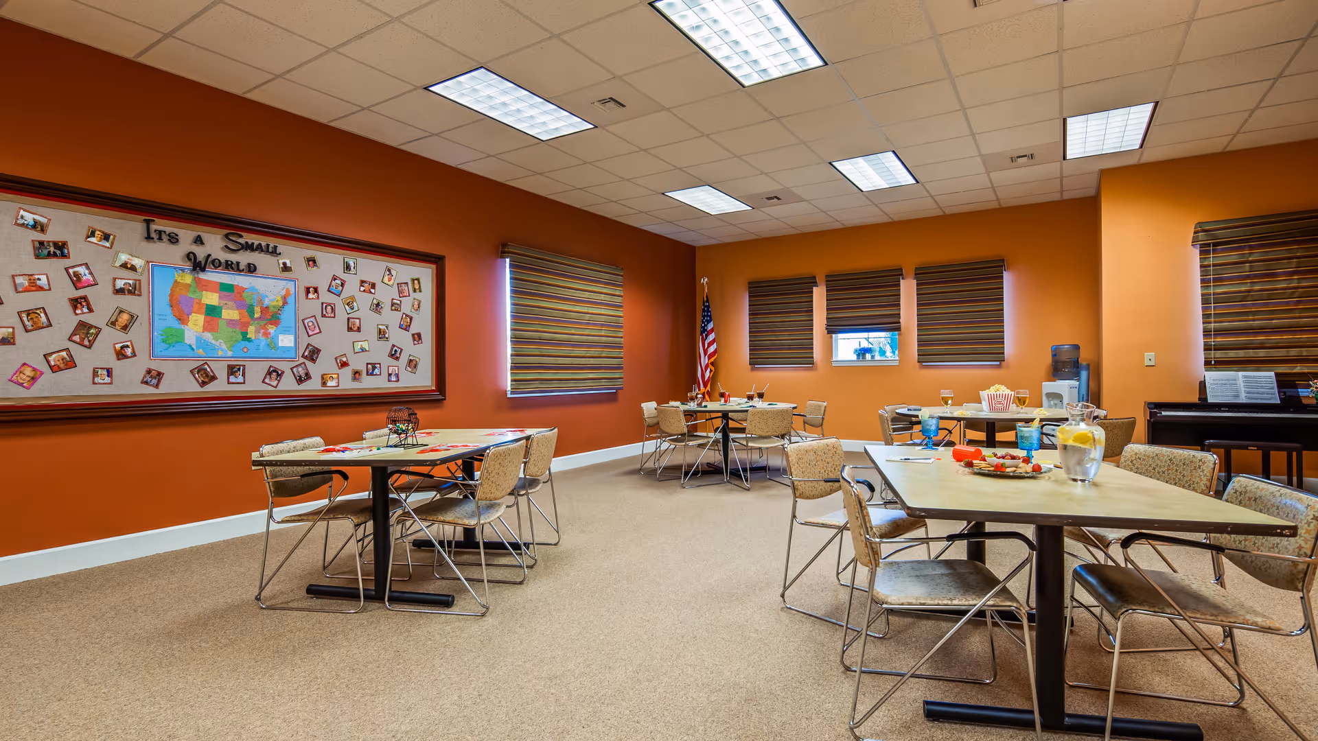 A community room with orange walls and beige carpet featuring several tables and chairs arranged for group activities. On the left wall, there is a large bulletin board with a map of the United States and photos surrounding it, titled 'It's a Small World.' The room has three windows with striped blinds, an American flag in the corner, a water dispenser, and a piano on the right side.