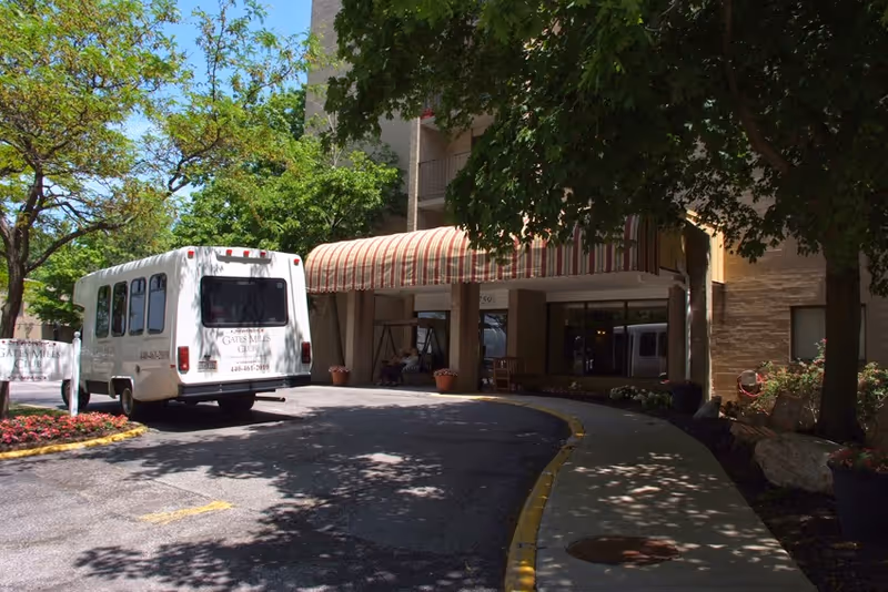 Entrance of Gates Mills Club Senior Living facility with a white shuttle bus parked near the covered driveway. The building entrance has a striped awning and is surrounded by trees and landscaped flower beds.