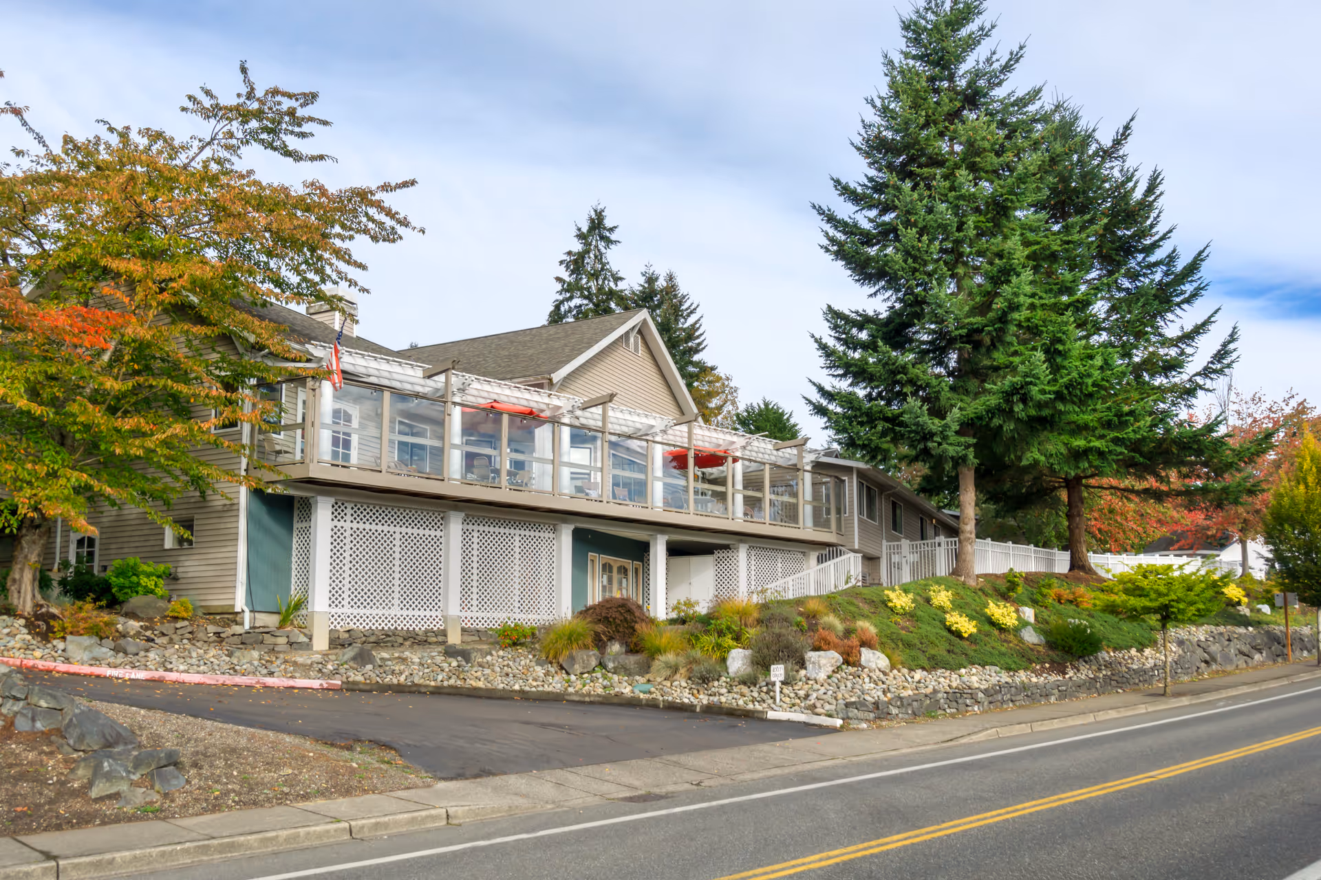 Exterior view of a senior living facility building with a raised deck featuring glass railings and outdoor seating. The building is surrounded by landscaped greenery, trees, and a stone retaining wall along a paved road.