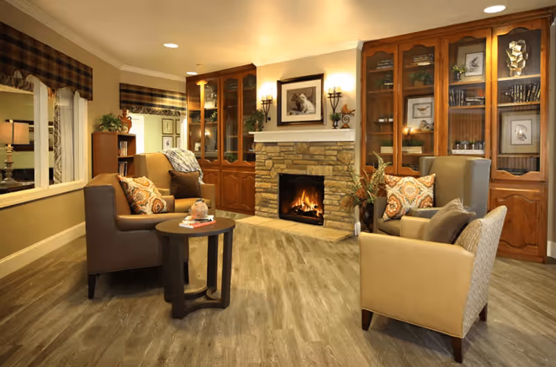 A cozy living room with a stone fireplace in the center, flanked by wooden cabinets with glass doors displaying books and decorative items. Four armchairs with patterned cushions are arranged around a small round wooden table. The room has wood flooring, warm lighting, and a window with curtains on the left side.