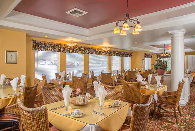 A bright and spacious dining room with multiple tables covered in yellow tablecloths and set with white napkins, cups, and silverware. The room features wicker chairs, large windows with blinds and floral valances, a decorative ceiling with a maroon inset, and elegant light fixtures. There are white columns and a large potted plant in the background.