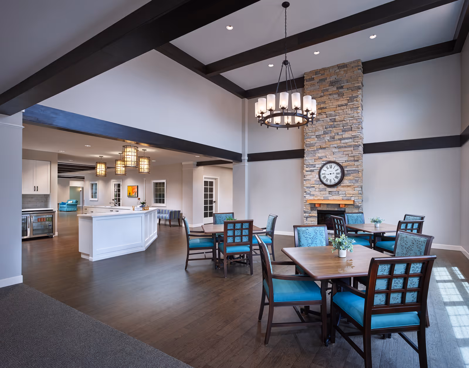 Bright communal dining area with wooden tables and turquoise upholstered chairs, a stone fireplace with a clock, and a white service island under exposed ceiling beams.