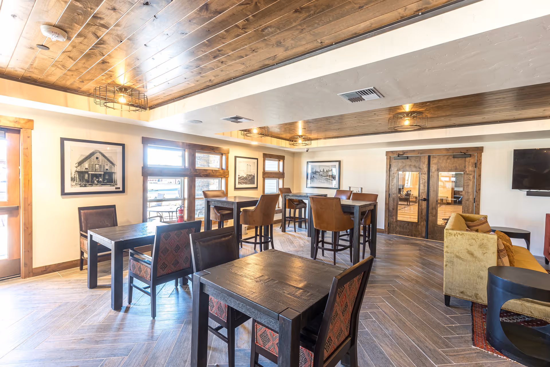 Interior view of a senior living facility lounge area with wooden tables and chairs, a mustard yellow couch, framed black and white photographs on the walls, wooden ceiling with modern light fixtures, and double wooden doors with glass panels.