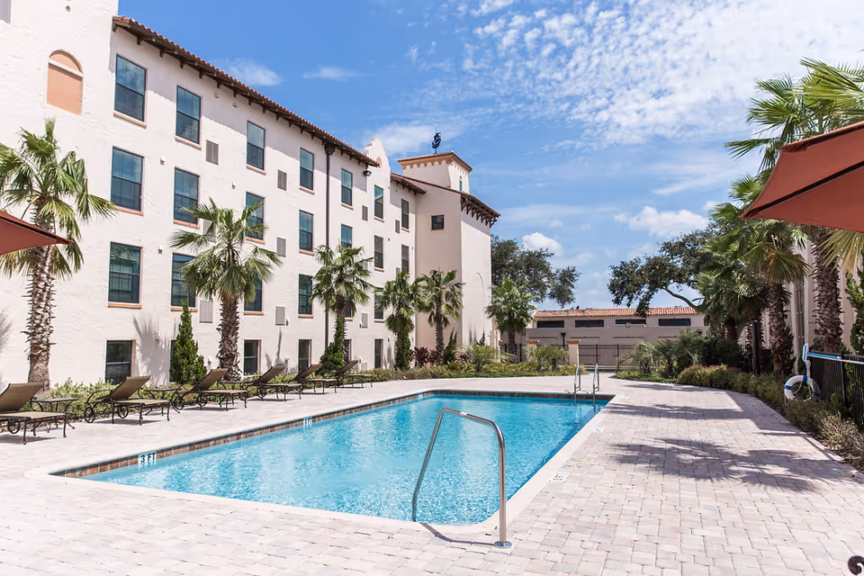 Outdoor swimming pool with lounge chairs, palm trees, and a multi-story Mediterranean-style building under a blue sky.