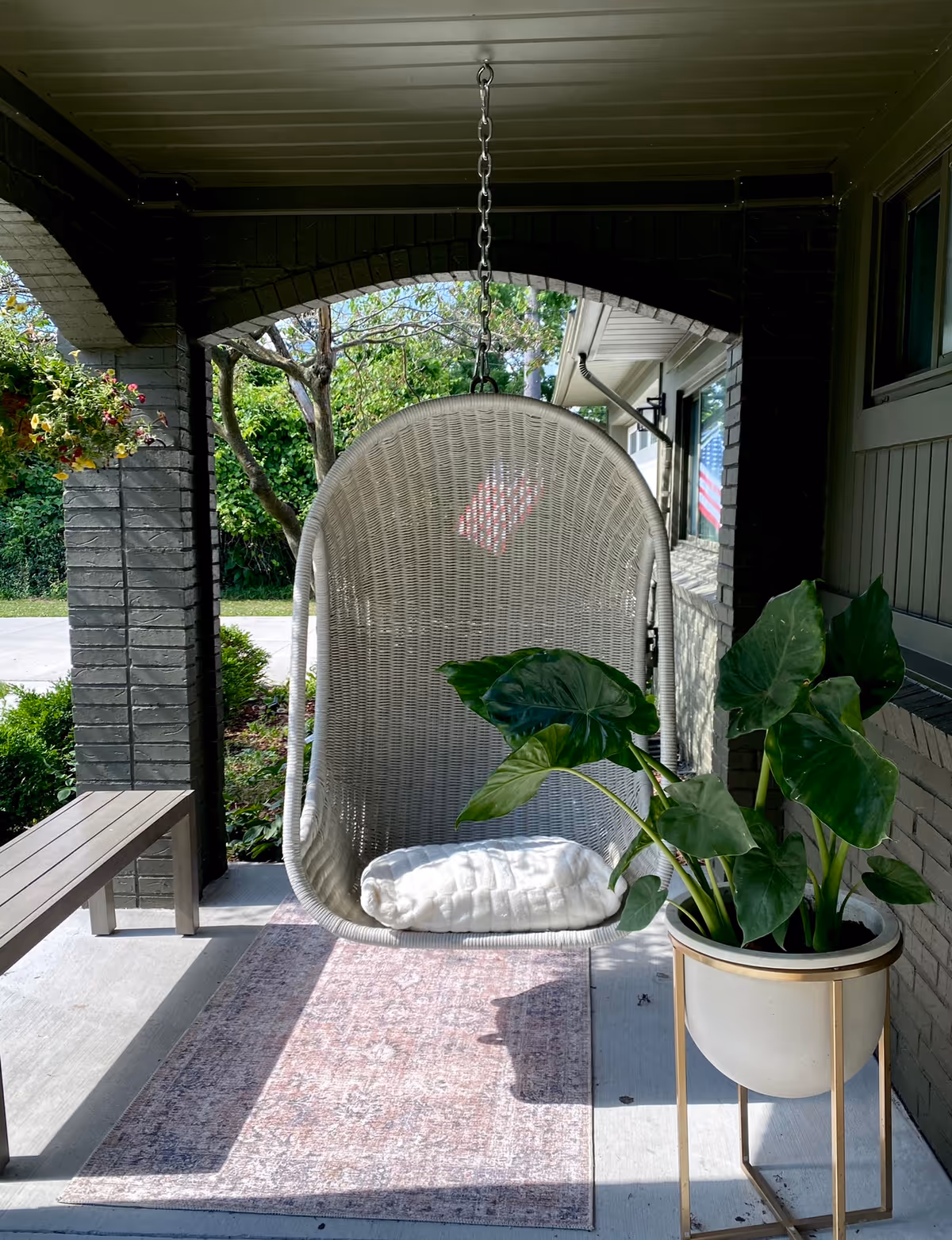 Covered front porch with a hanging wicker swing chair, potted plant, bench, and rug.