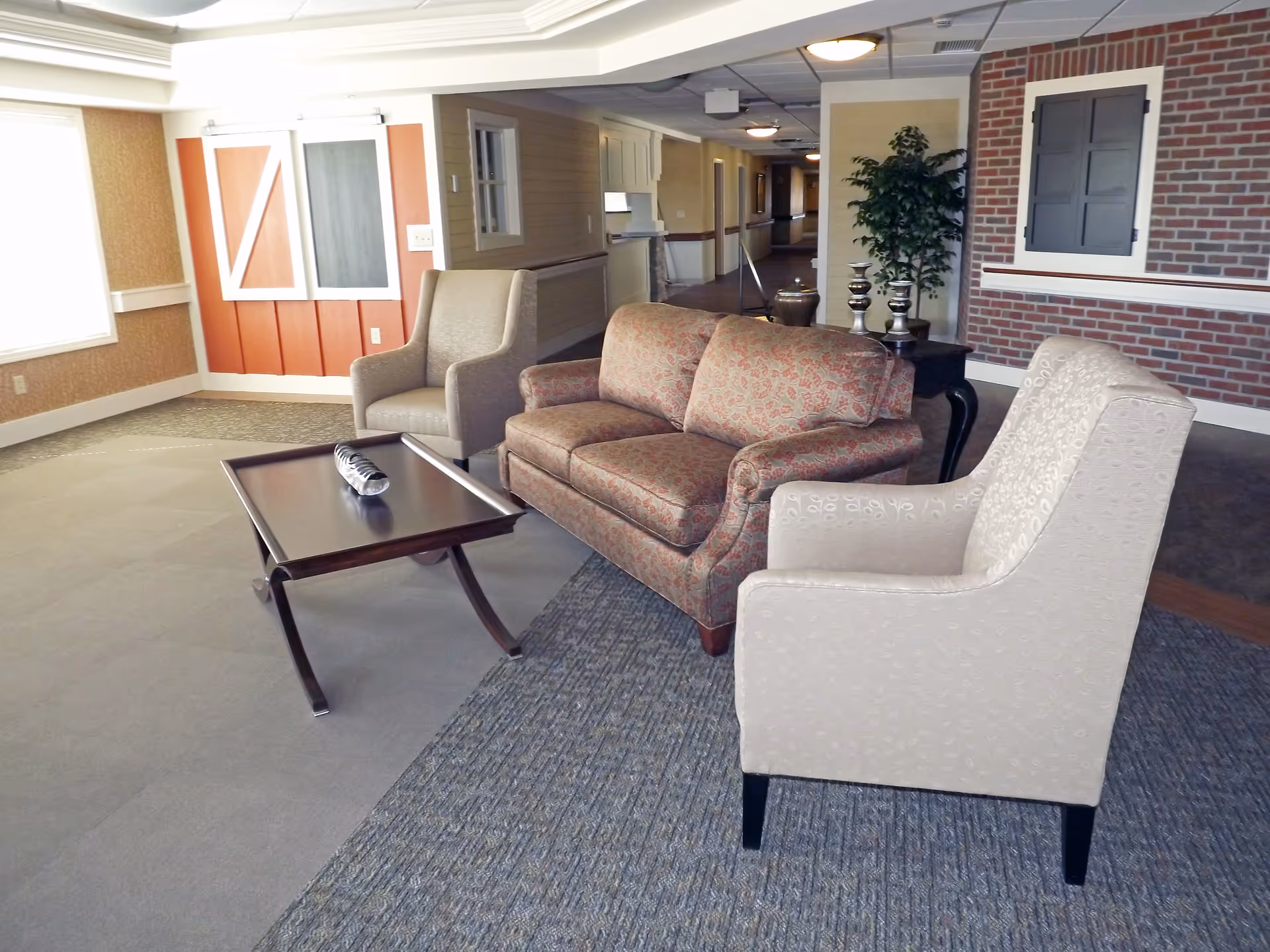 A cozy seating area in a senior living facility featuring a patterned loveseat, two beige armchairs, and a dark wooden coffee table with a decorative item on it. The room has a mix of carpet and tile flooring, a brick accent wall with a closed window shutter, and a potted plant on a side table. The background shows a hallway with soft lighting and neutral-colored walls.