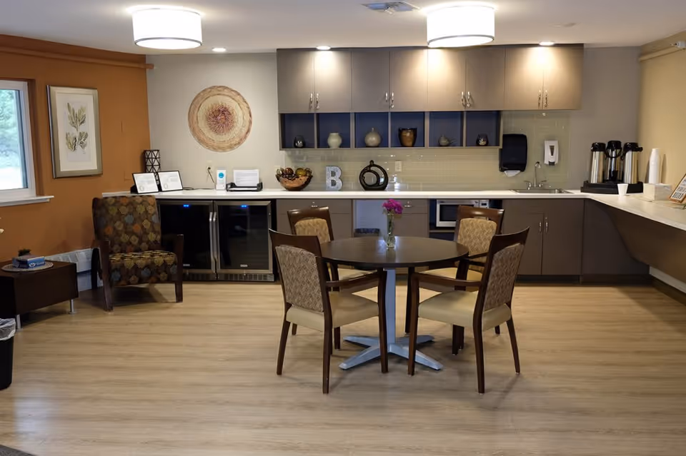 A cozy common area with a round table and four chairs in the center. Behind the table is a kitchenette with cabinets, a sink, a microwave, and coffee dispensers. To the left, there is a patterned armchair next to a window and wall art. The room has wood flooring and soft lighting from ceiling fixtures.