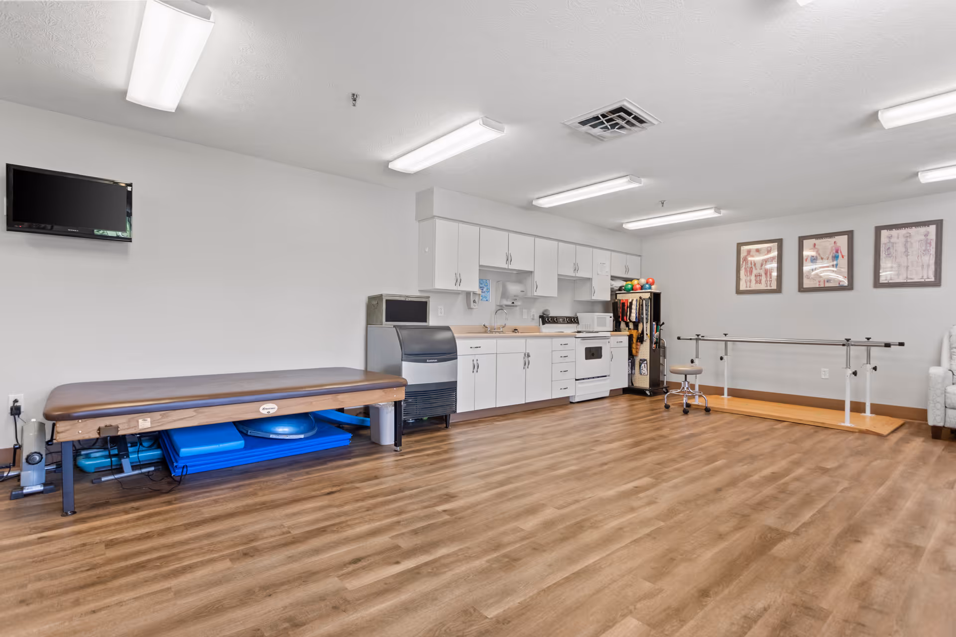 A spacious room with wood flooring featuring a therapy table, exercise equipment, a kitchenette with white cabinets, a stove, microwave, and refrigerator. There are parallel bars for physical therapy, a rolling stool, and framed anatomical charts on the wall. The room is well-lit with ceiling lights.