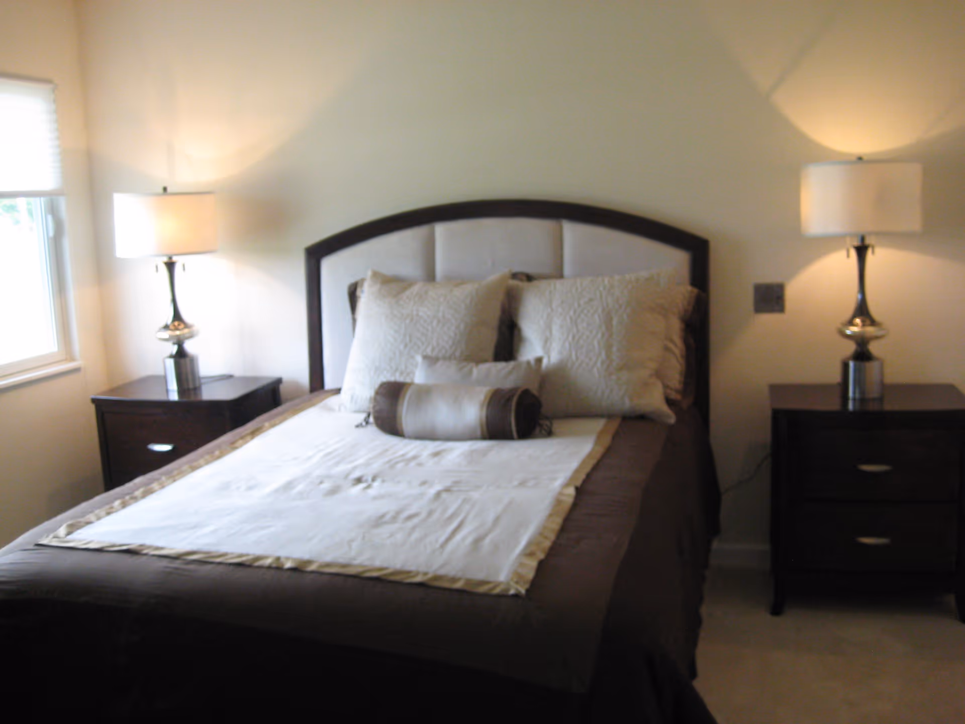 A neatly made bed with a padded headboard, two large pillows, and a cylindrical bolster pillow in a bedroom. There are two matching dark wood nightstands on either side of the bed, each with a silver lamp with a white lampshade. A window with blinds is visible on the left side of the room.