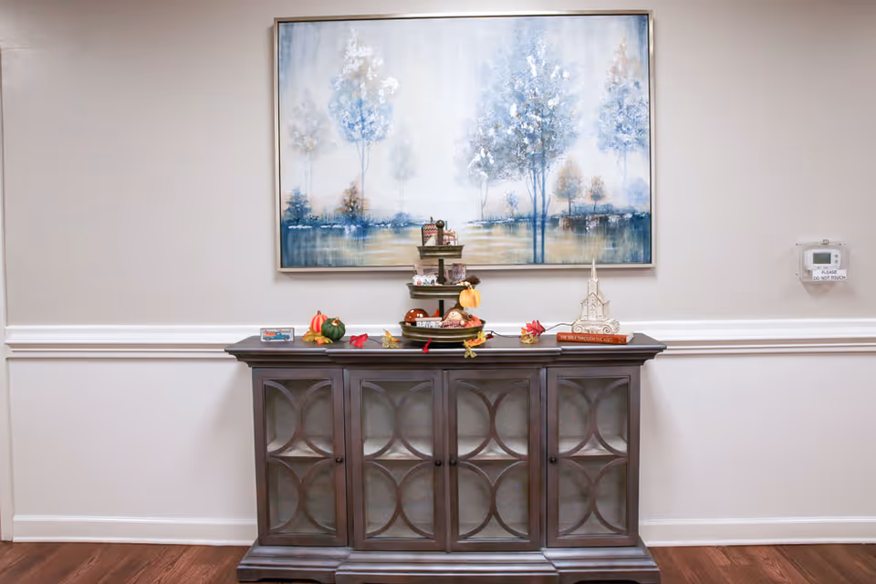 A decorative wooden cabinet with glass-paneled doors stands against a beige wall with white trim. On top of the cabinet are small seasonal decorations including mini pumpkins, leaves, and a tiered tray with various items. Above the cabinet hangs a large framed painting depicting a serene landscape with trees in soft blue and neutral tones. A thermostat with a 'Please Do Not Touch' sign is mounted on the wall to the right.