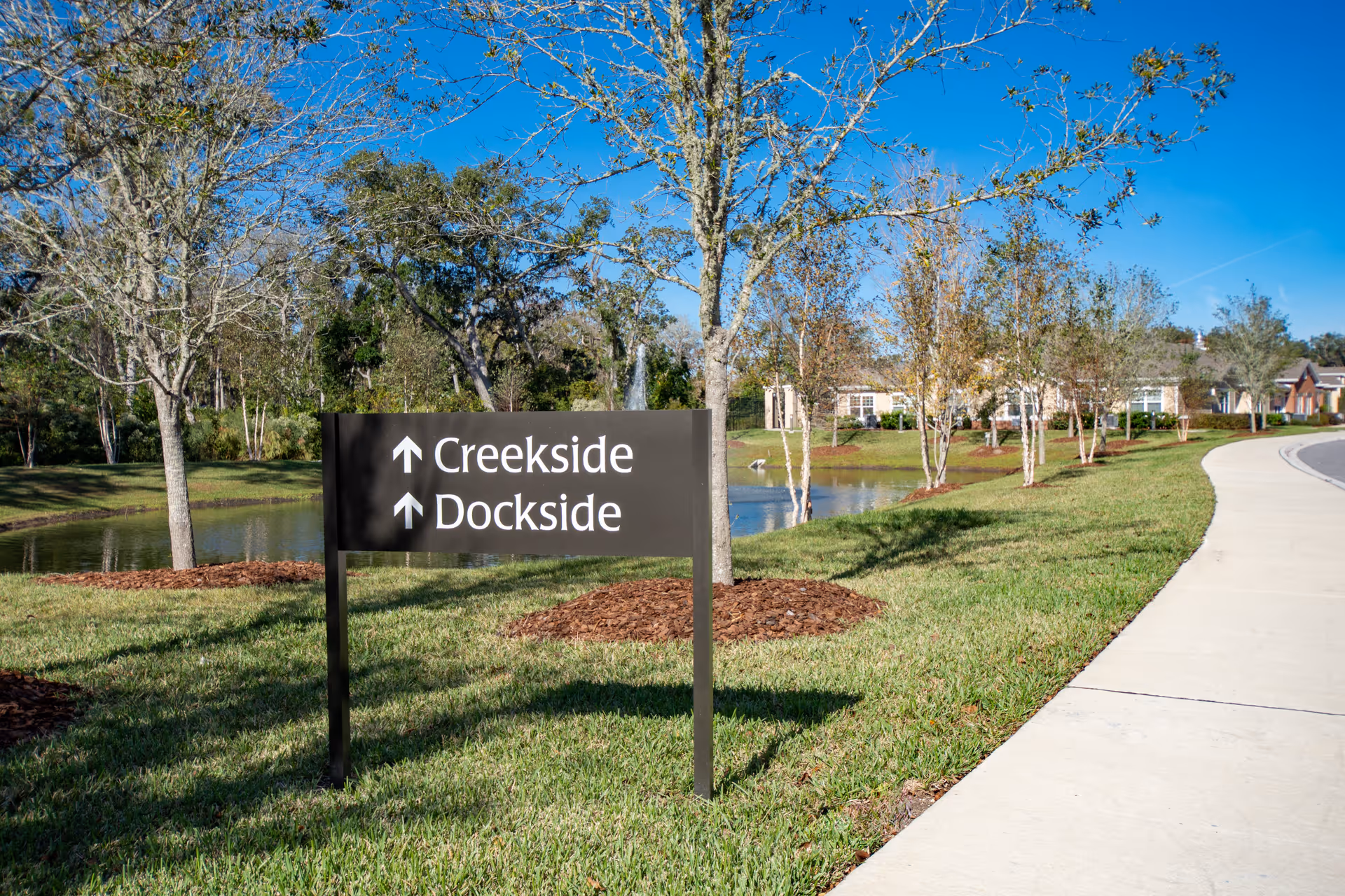 Outdoor scene at Anthem Lakes featuring a black directional sign with white text pointing towards Creekside and Dockside. The sign is placed on a grassy area with trees, a pond, and a clear blue sky in the background. A curved sidewalk runs along the right side of the image.