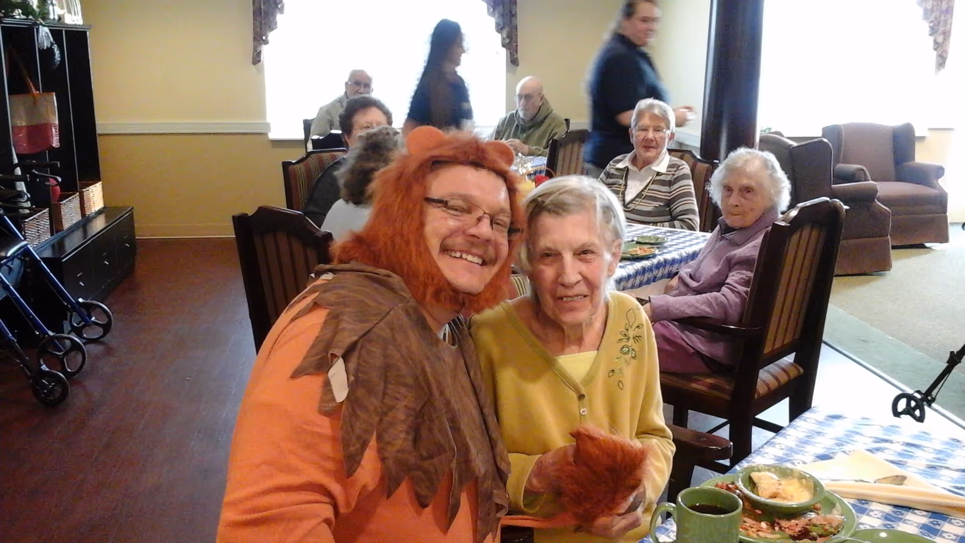 A group of elderly people sitting around tables in a dining area. In the foreground, a man dressed in a lion costume with a mane and glasses is smiling and posing with an elderly woman wearing a yellow sweater. Other elderly individuals are seated at tables in the background, with plates of food and cups visible on the tables. The room has wooden flooring and light-colored walls with windows letting in natural light.