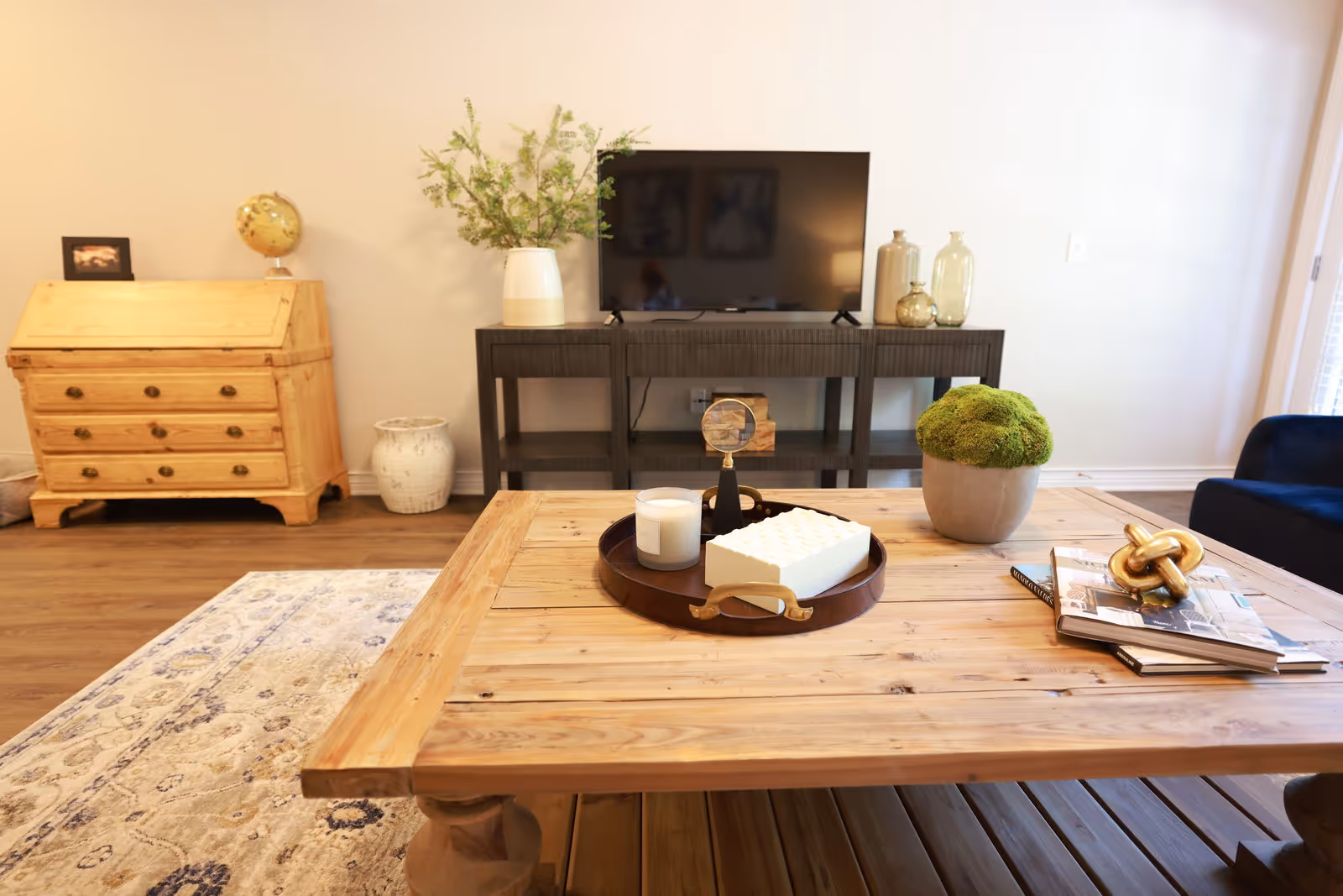 A cozy living room with a wooden coffee table in the foreground decorated with a tray holding a candle, a white decorative item, and a magnifying glass. There is also a potted green plant and some books with a golden knot sculpture on the table. In the background, a dark wooden TV stand holds a flat-screen TV and decorative vases, while a light wooden chest of drawers with a globe and framed photo sits against the wall. The room has a light-colored rug and wooden flooring.