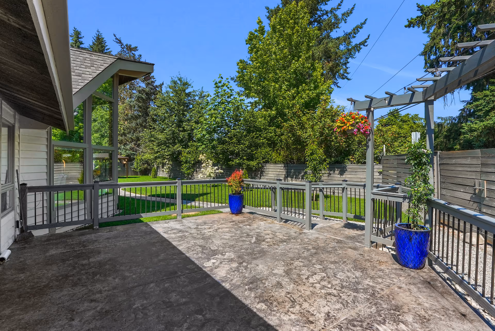 A spacious outdoor patio area with a concrete floor, gray railing, and a pergola structure. Two large blue pots with plants are placed on the patio. The patio overlooks a green lawn with trees and a wooden fence under a clear blue sky.