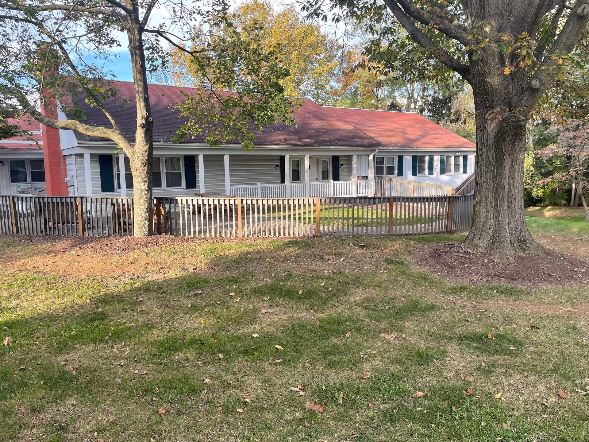 Exterior view of a single-story building with a red roof and white siding, surrounded by a wooden fence and large trees with autumn leaves. The building has multiple windows with green shutters and a small porch area.