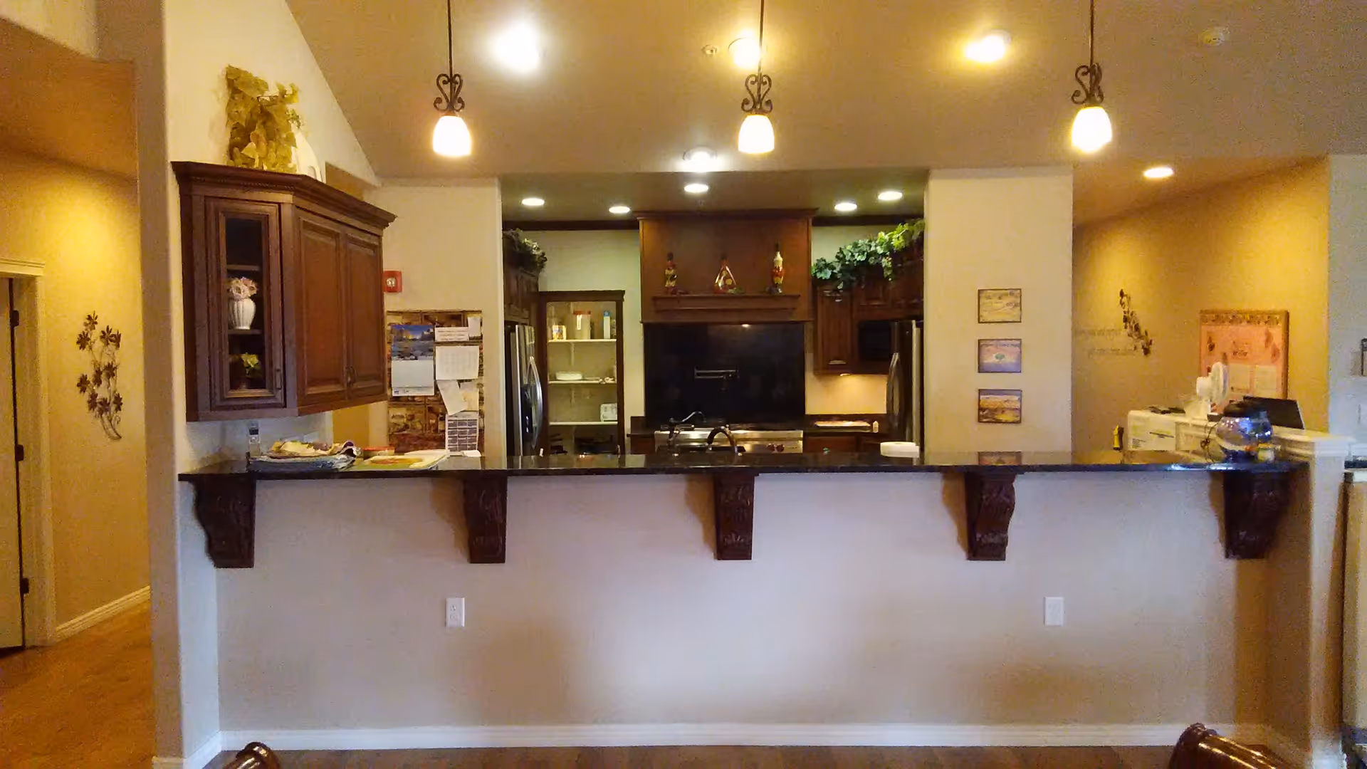 Interior view of a kitchen area in an assisted living facility featuring a dark granite countertop bar with decorative wooden corbels, wooden cabinets, stainless steel appliances, and warm lighting from ceiling fixtures. There are decorative plants and items on top of the cabinets and a bulletin board on the right wall.