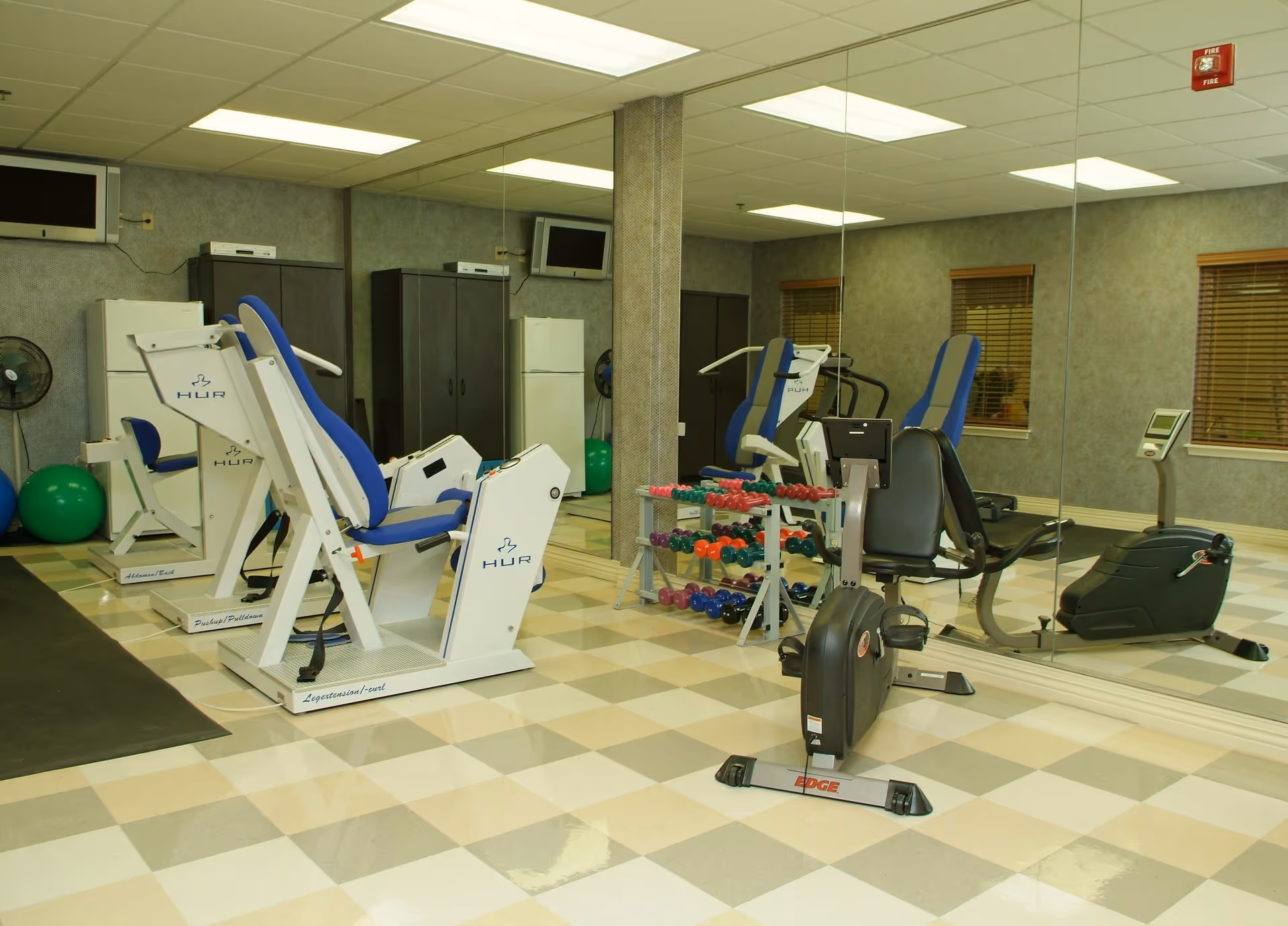 A fitness room in a senior living community featuring exercise machines with blue seats, a stationary bike, a rack of colorful dumbbells, large mirrors on the wall, and a checkered tile floor.