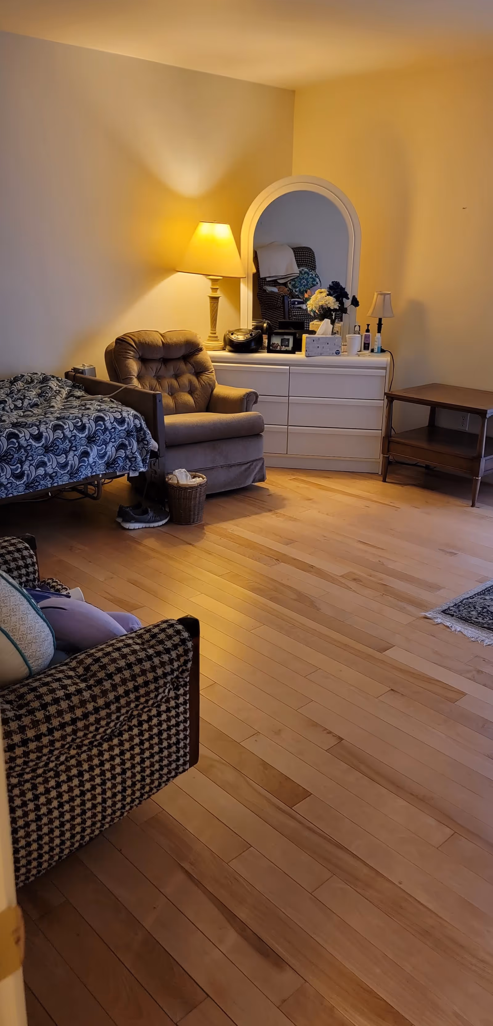 A cozy room with wooden flooring featuring a bed with a patterned blue and white blanket, a cushioned armchair, a white dresser with an arched mirror, a table lamp, and various personal items including a radio, framed photo, flowers, and hand sanitizer. There is also a small wooden side table and part of another cushioned chair with a houndstooth pattern visible in the foreground.