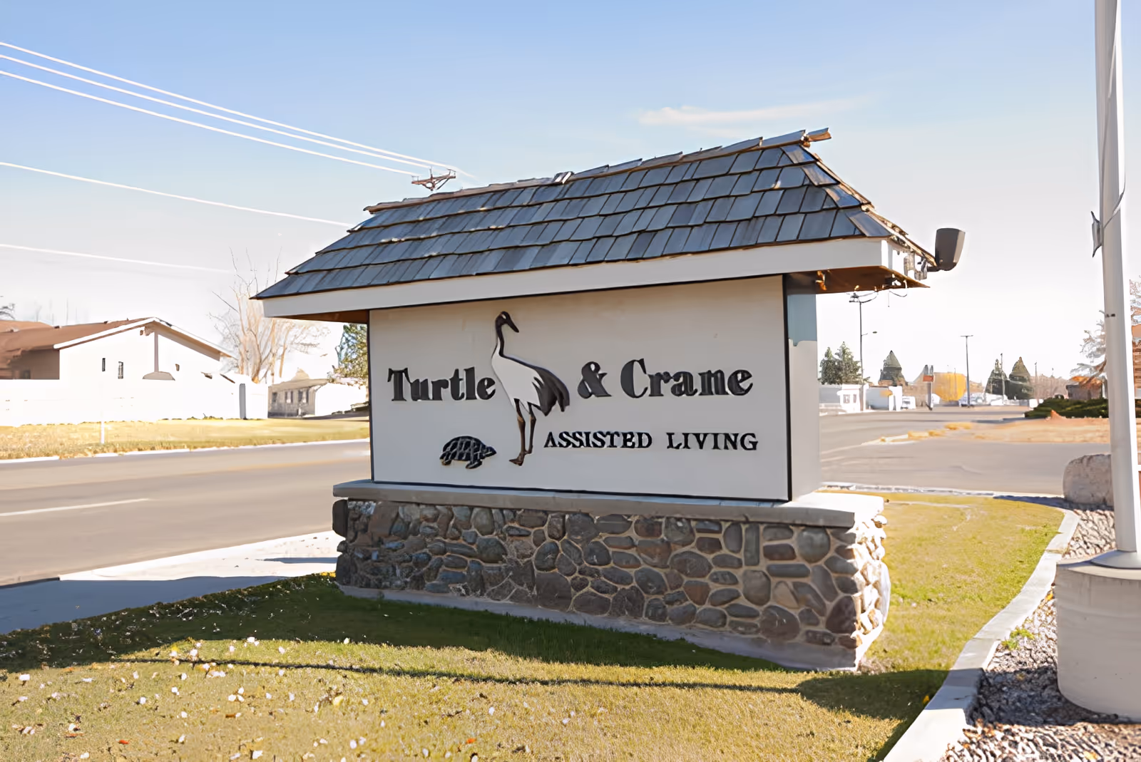 Outdoor sign for Turtle & Crane Assisted Living with a stone base and a shingled roof, located near a street with houses and trees in the background.
