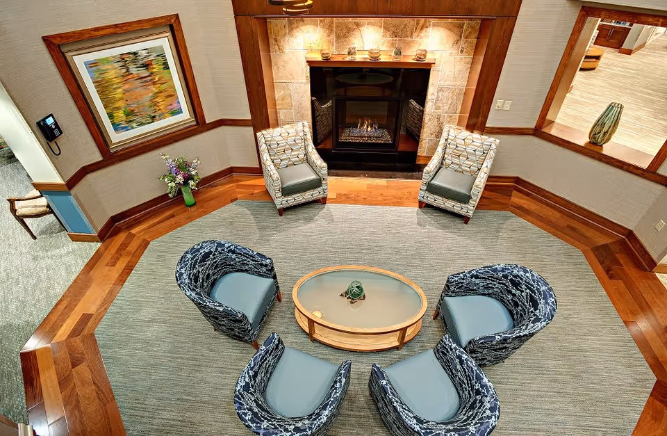 A cozy seating area with a fireplace, featuring two patterned armchairs and four blue cushioned chairs arranged around an oval glass coffee table on a large rug. The room has wooden flooring and walls with a framed abstract painting and a vase of flowers. A window opens to an adjacent room with a decorative vase on a counter.