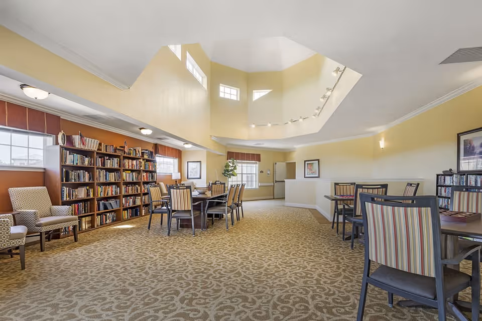 A spacious, well-lit common area with high ceilings and multiple windows. The room features several tables and chairs arranged for group seating, a large bookshelf filled with books, and comfortable armchairs along the wall. The walls are painted in warm tones, and framed artwork is displayed on the walls.