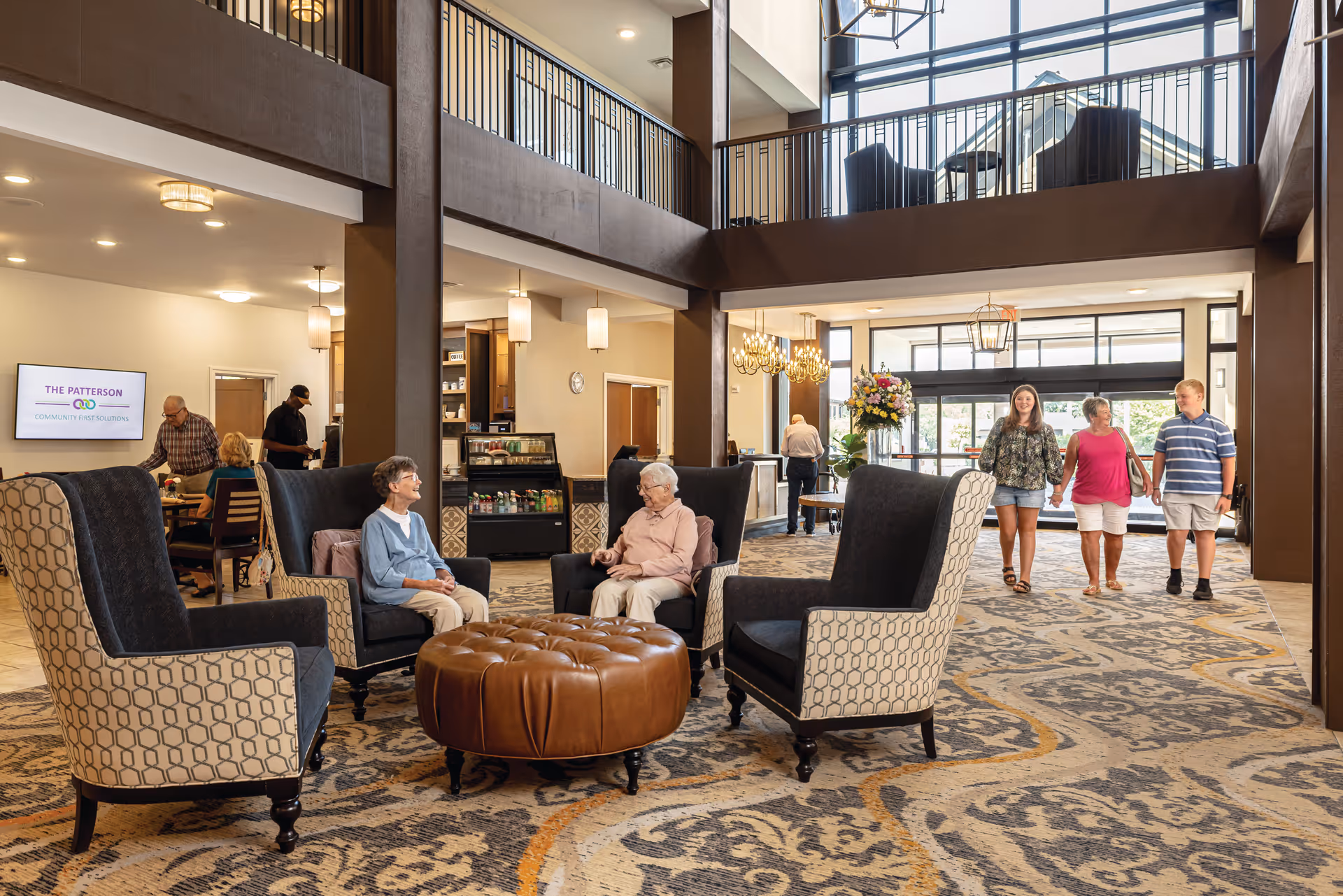 A spacious and well-lit senior living facility lobby with high ceilings and large windows. Four elderly women are seated in comfortable armchairs arranged around a round leather ottoman, engaged in conversation. In the background, a few people are walking and sitting at tables near a wall-mounted screen displaying 'The Patterson'. The area features patterned carpeting, modern lighting fixtures, and a flower arrangement near the entrance.