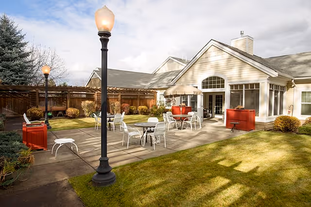 Outdoor patio area at Brookdale Nine Mile with several white metal tables and chairs, a red umbrella, two black lamp posts, and a beige building with large windows and a chimney in the background. The area is surrounded by a wooden fence and landscaped with bushes and grass.