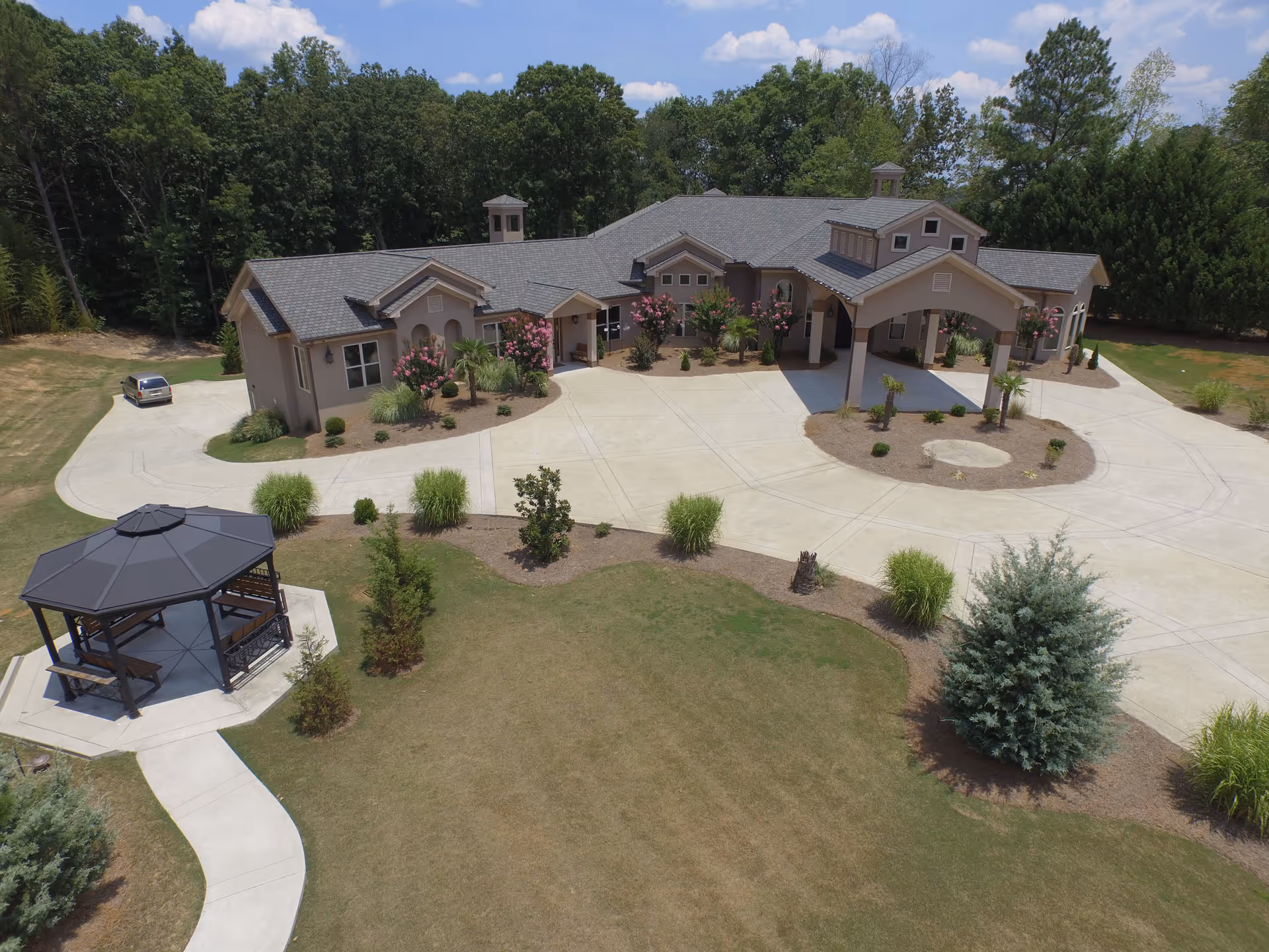 Aerial view of a single-story senior living facility building with a large driveway and entrance canopy. The building is surrounded by landscaped gardens with bushes, small trees, and flowering plants. A gazebo with benches is visible on the left side, connected by a paved walkway. The background includes a wooded area under a partly cloudy sky.