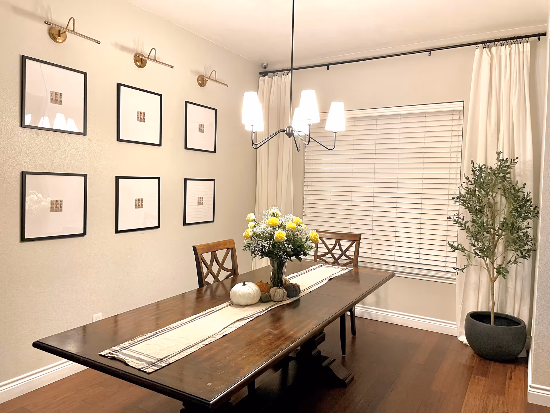 A dining room with a long wooden table set with a white and beige table runner, a vase of yellow and white flowers, and decorative pumpkins. Two wooden chairs are positioned at the table. The room has six framed pictures on the wall, a window with closed blinds and white curtains, a hanging light fixture with five lamps, and a potted plant in the corner.