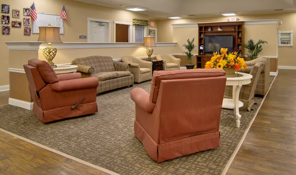 A cozy senior living lounge with upholstered armchairs and sofas arranged around a table and a television cabinet.