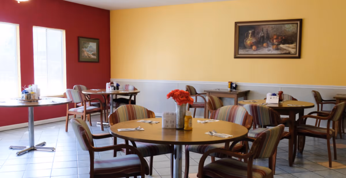 A dining room with round wooden tables and striped cushioned chairs. Each table is set with utensils and condiments, and there are paintings on the red and yellow walls. Large windows allow natural light to brighten the room.