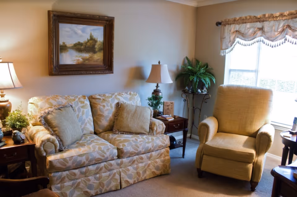 A cozy living room area with a patterned loveseat adorned with two beige cushions, a beige armchair, two wooden side tables with lamps and plants, a framed landscape painting on the wall, and a window with a decorative valance letting in natural light.