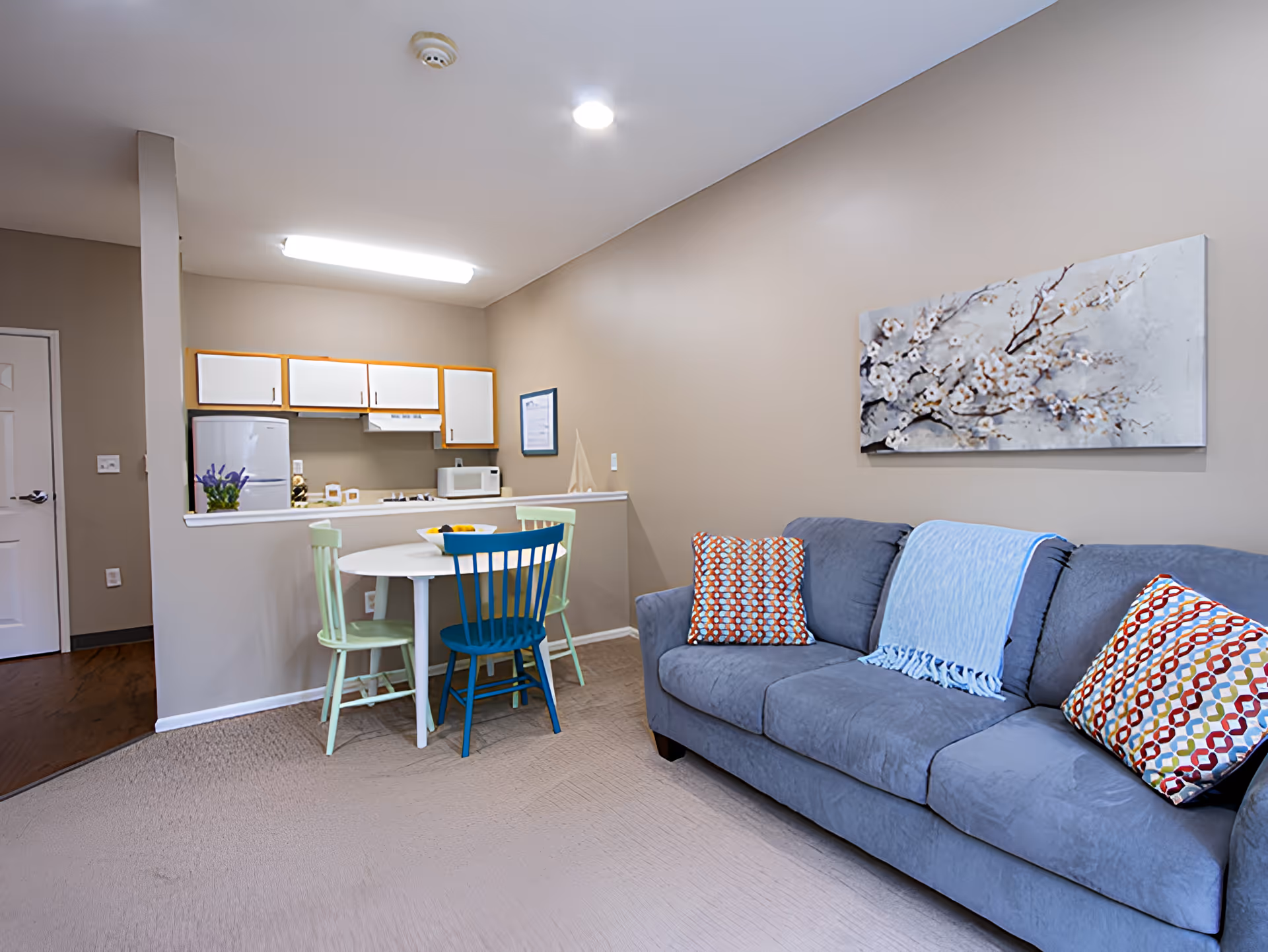 A cozy living area with a gray sofa adorned with colorful patterned pillows and a light blue throw blanket. On the wall above the sofa is a painting of white blossoms on branches. Adjacent to the living area is a small dining space with a round white table and three chairs in pastel green and blue. Behind the dining area is a compact kitchen with white cabinets, a refrigerator, microwave, and stove. The walls are painted beige and the floor is carpeted in the living and dining area with wood flooring near the kitchen entrance.