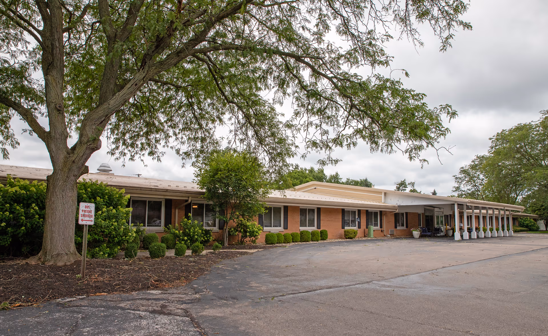 Single-story brick senior living building with a covered entrance, a large tree and shrubs in front, and an empty paved driveway.