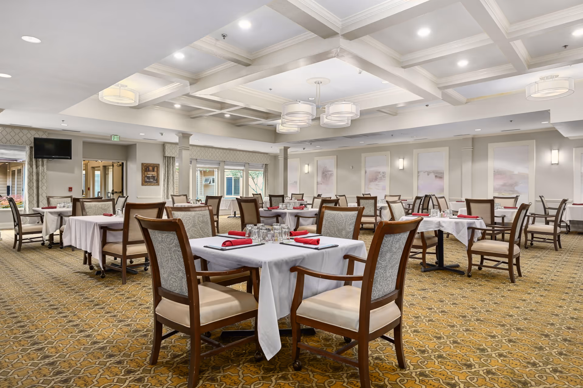 A spacious dining room with multiple tables covered in white tablecloths, each set with glasses and red napkins. The room features patterned carpet, cushioned wooden chairs, soft lighting from ceiling fixtures, and large windows letting in natural light.