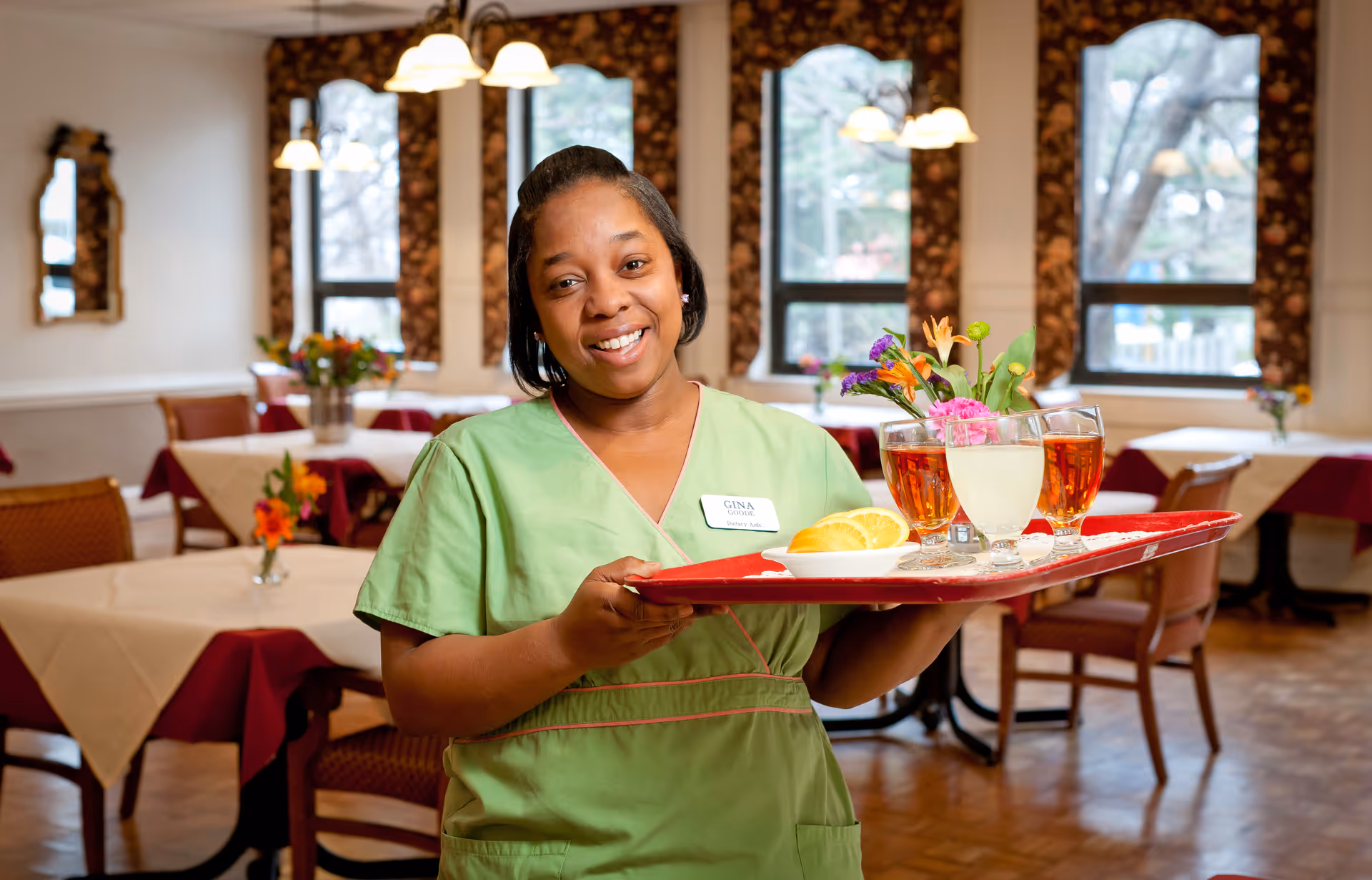 A smiling woman wearing a green uniform and a name tag that reads 'Gina' is holding a red tray with drinks, a plate of bread, and a small flower arrangement in a dining room with tables covered in white and burgundy tablecloths and large windows in the background.