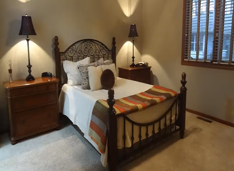 A cozy bedroom with a wooden bed frame featuring an ornate metal headboard. The bed is made with white sheets, multiple decorative pillows, and a colorful striped blanket at the foot. There are two wooden nightstands on either side of the bed, each with a dark lamp. A window with wooden blinds is on the right wall, and the room has beige walls and carpeted floor.