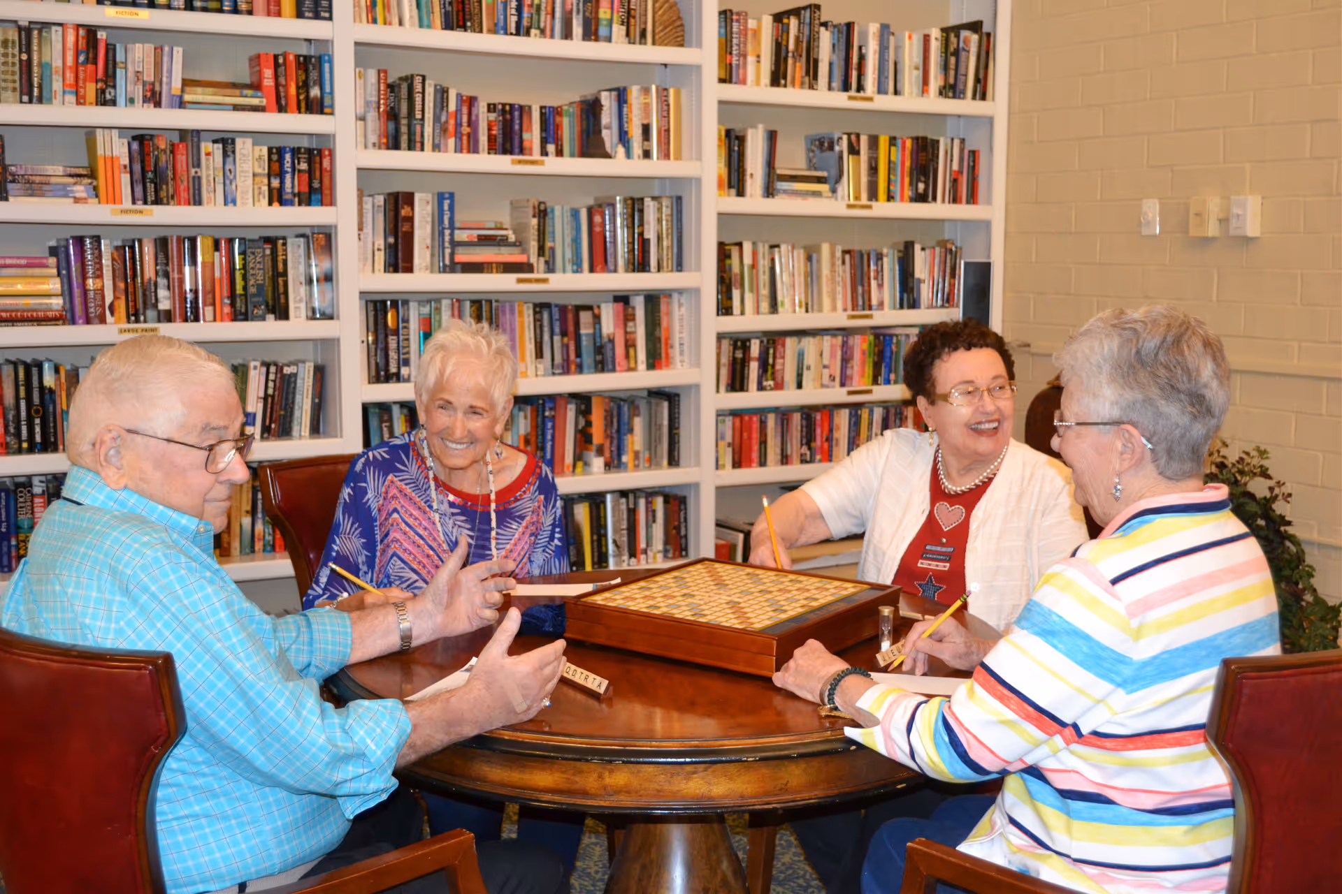 Four elderly people sitting around a round wooden table playing a board game in a room with bookshelves filled with books in the background. They appear to be engaged and smiling, enjoying their time together.